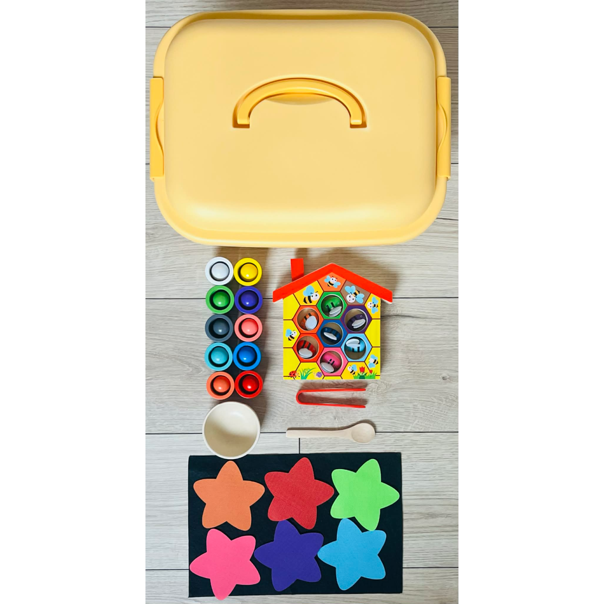 Picture shows yellow bin, wooden colorful bee hive with little bees inside, a red tweezer, a bowl, a wooden spoon, wooden cups and balls of solid rainbow colors, 6 colorful velcro stars, and a black felt sheet. 