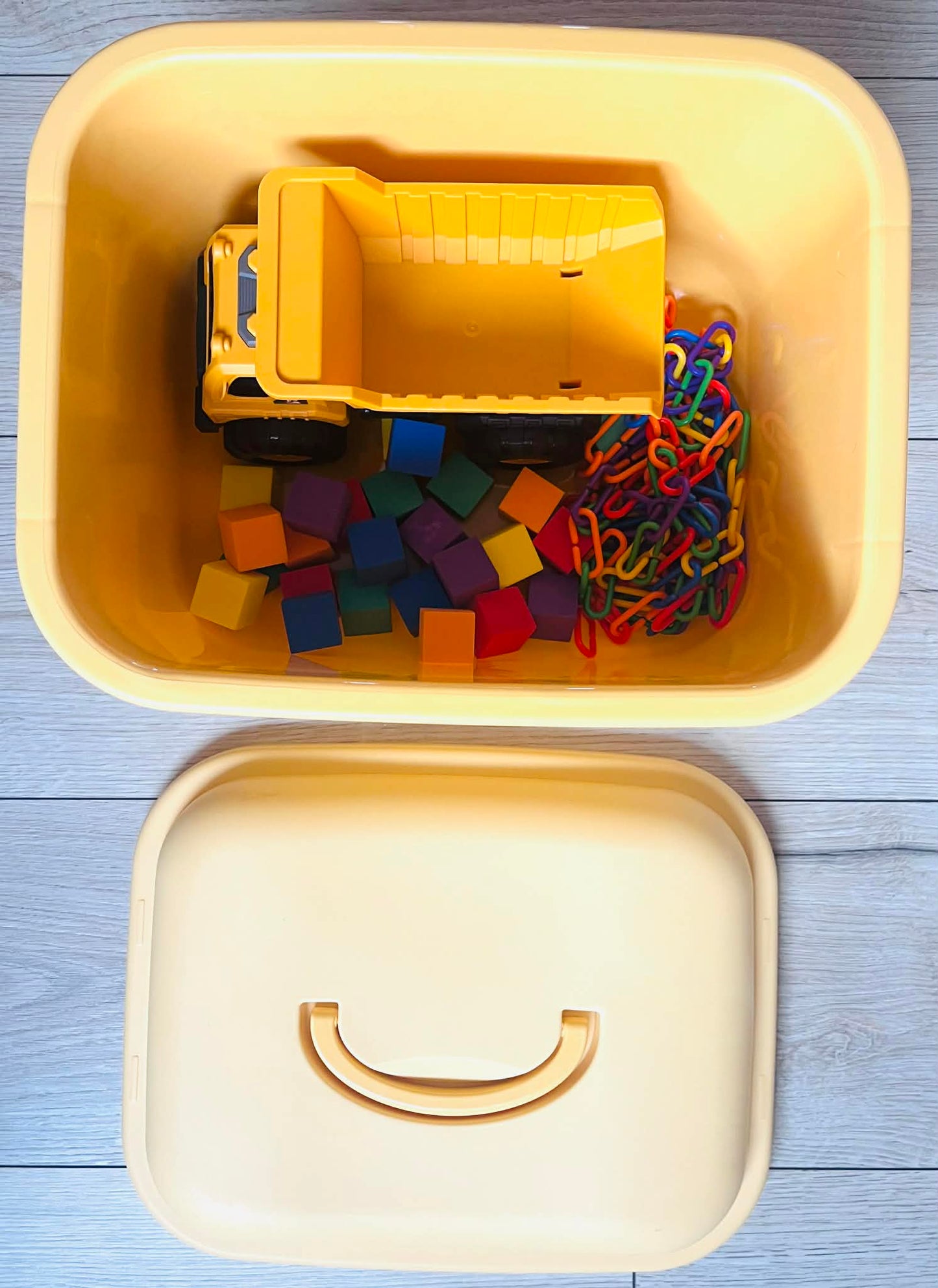Yellow toy truck and colorful building blocks and links in a yellow container on a light wooden surface.