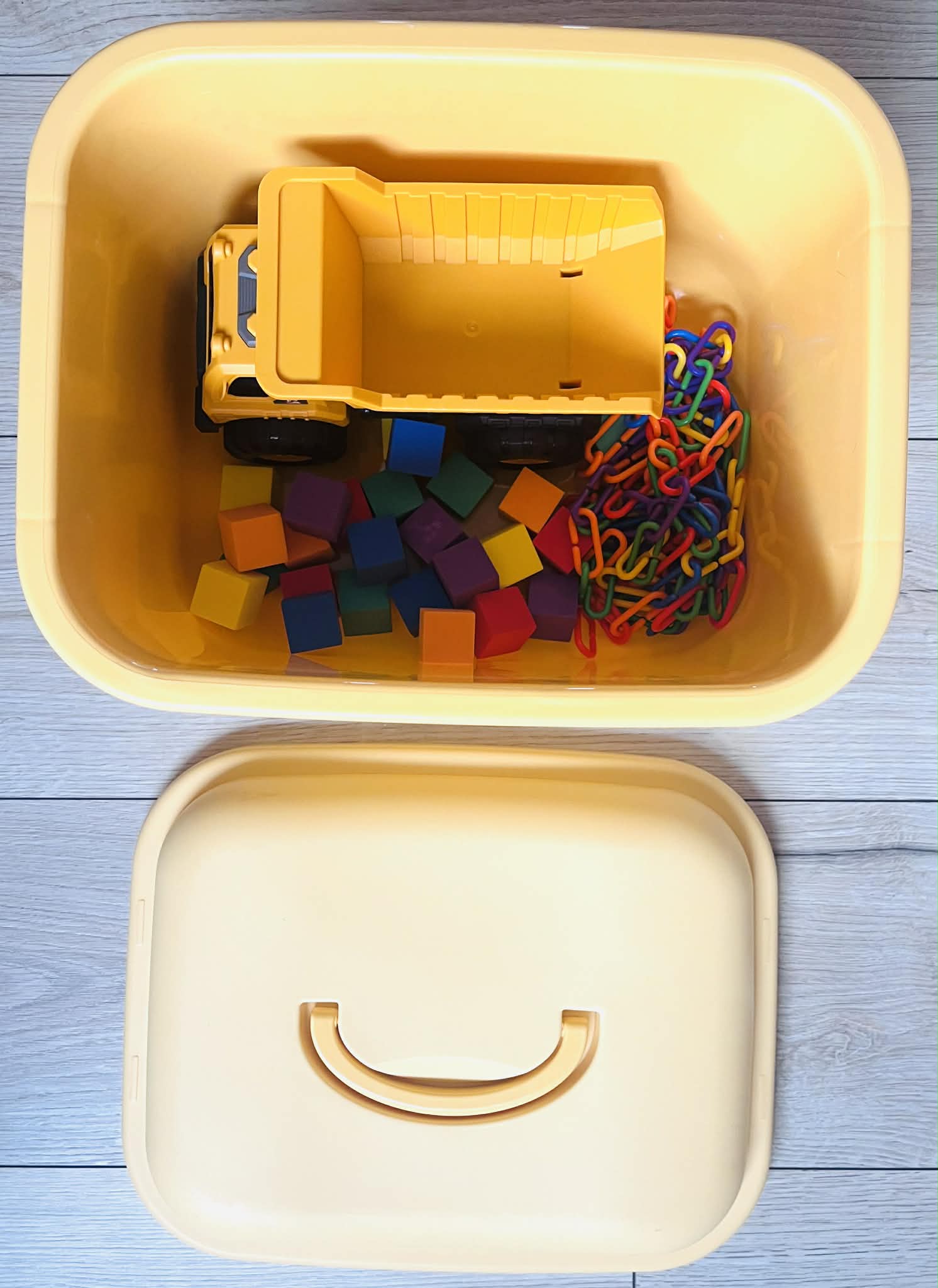 Yellow toy truck and colorful building blocks and links in a yellow container on a light wooden surface.