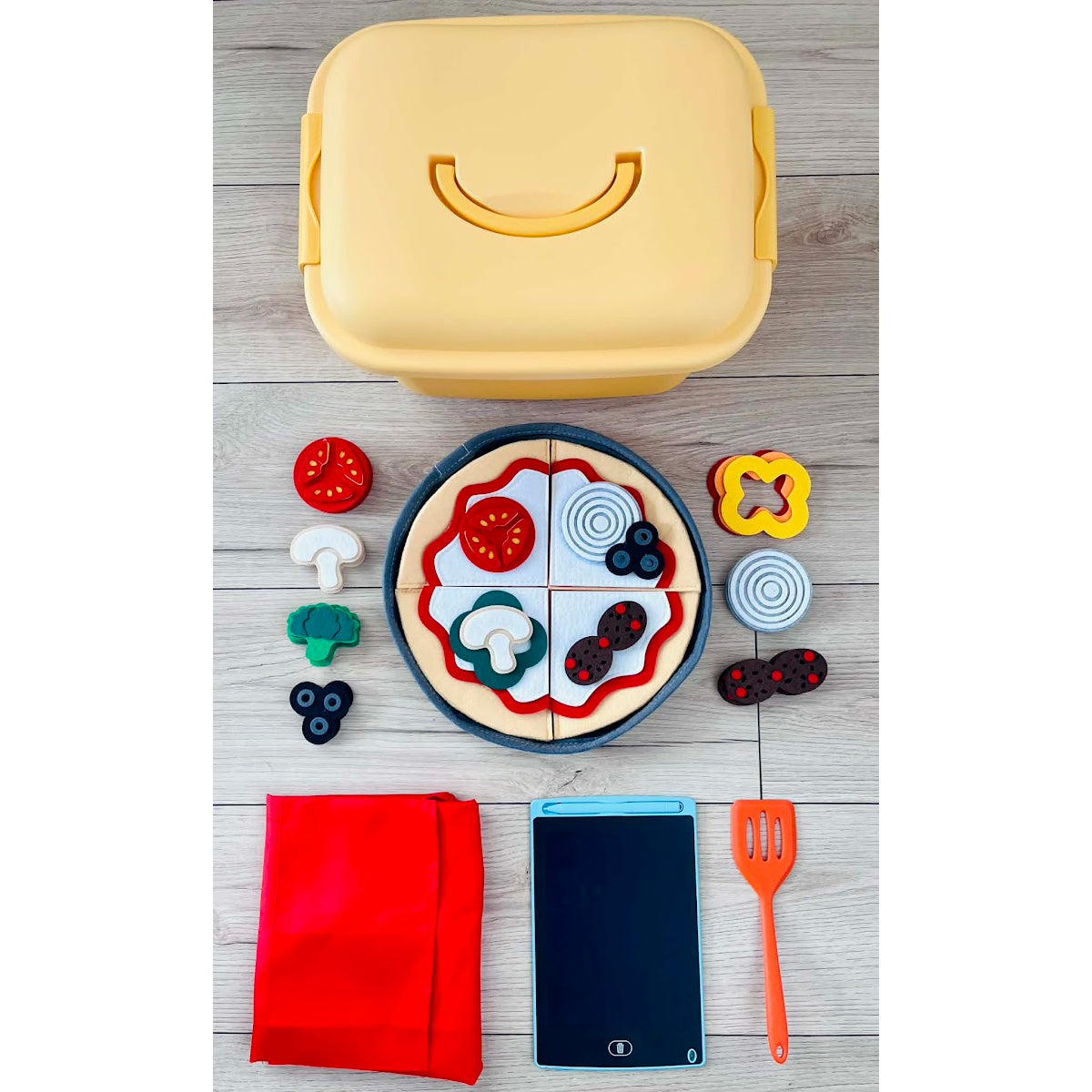 Yellow plastic bin with felt pizza-themed cutouts, red apron, tablet, and orange spatula on a wooden surface.